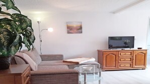 A living room with a sofa, a coffee table, a TV on a wooden cabinet, a painting on the wall, and a large plant. - Vacation home Figelski with terrace in Sassnitz - Holiday home Figelski in Sassnitz (Sassnitz auf Rügen)