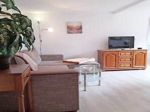Ein Wohnzimmer mit einer Couch, einem Couchtisch, einem Fernseher auf einer Holzkommode, einem Bild an der Wand und einer großen Pflanze.