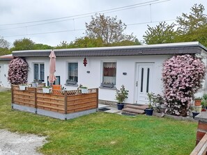 Ein eingeschossiges Haus mit Veranda, einer kleinen Terrasse und einem Gartenbereich mit Kübelpflanzen.