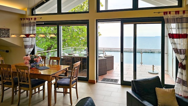Uma área de refeições com mesa e cadeiras, um sofá e vista para o mar por grandes janelas.