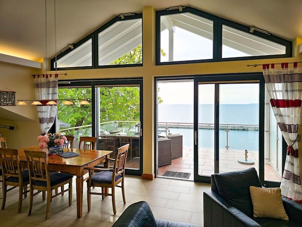 A dining area with a table and chairs, a sofa, and a view of the sea through large windows. - Villa Friede-Marie, Penthouse Lenea - magnificent sea view over the Baltic Sea - (Sassnitz auf Rügen)