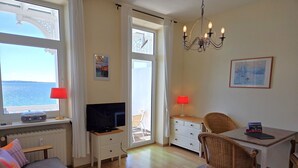 Una habitación con televisor, una mesa de comedor con sillas, un sofá, una ventana con vista al mar y un candelabro.
