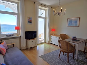 Ein Zimmer mit Fernseher, Esstisch mit Stühlen, einer Sitzecke und einem Fenster mit Meerblick, sowie einer Kristallleuchter.