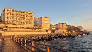 Un paseo marítimo con un camino de madera, edificios con ventanales de arco y una costa rocosa.