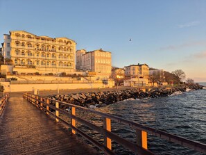 Eine Uferpromenade mit Holzsteg, Gebäuden mit Rundbogenfenstern und einer felsigen Küstenlinie.