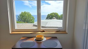 Et værelse med et stort vindue der udover en strand, et lille bord med en skål, og trægulv.