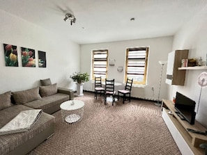 Ein Wohnzimmer mit einer Couch, einem Couchtisch, zwei Sesseln, einem Fernseher auf einem Schrank und einer Pflanze.
