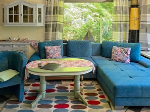 Ein Wohnzimmer mit blauem Sofa, rundem Tisch, einer Kommode und Blick ins Grüne.