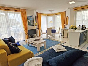 A hotel room with a sofa, a dining table, a TV, a kitchen area, and large windows with curtains. - Vacation apartment Anemone - vacation with dog on the edge of the national park (Sassnitz auf Rügen)