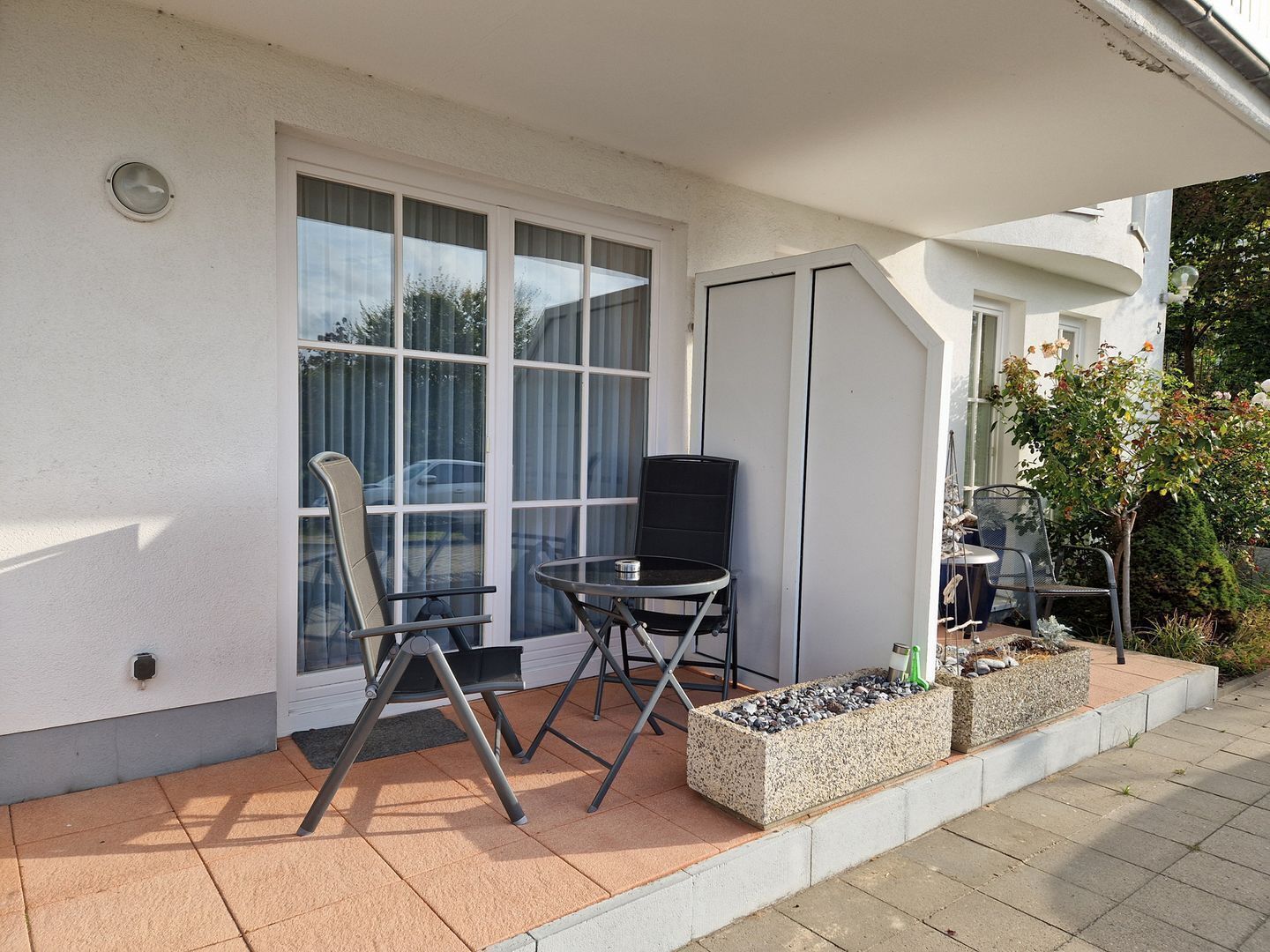 A patio with a table and chairs, a small garden bed, and a white building with large windows.