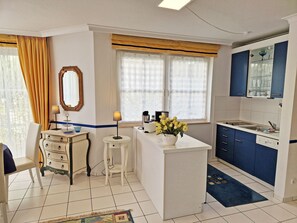 A compact kitchen with a central island, a dining area with a chair, and a window with curtains. - Vacation apartment Anemone - vacation with dog on the edge of the national park (Sassnitz auf Rügen)