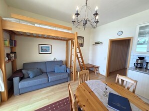 Ein kompakter Wohnraum mit Sofa, Esstisch, einer Leitern, einem Bücherregal und einer Küchenzeile.