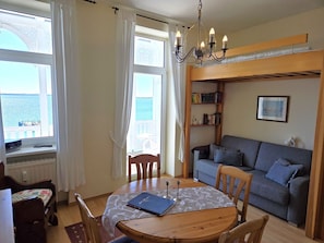 Ein gemütliches Zimmer mit einem runden Holztisch, einem grauen Sofa, einem Bücherregal und Meerblick durch große Fenster.