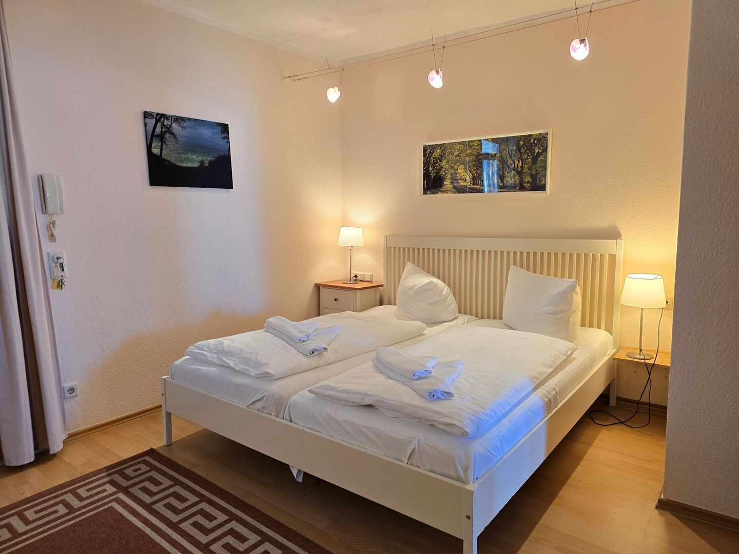 Un lit double avec du linge de lit blanc, deux tables de chevet avec des lampes, une petite table et deux tableaux accrochés au mur.