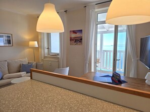 Une chambre d’hôtel moderne avec un canapé, une table d’appoint en bois, une lampe, une fenêtre avec des rideaux et une vue sur la mer.