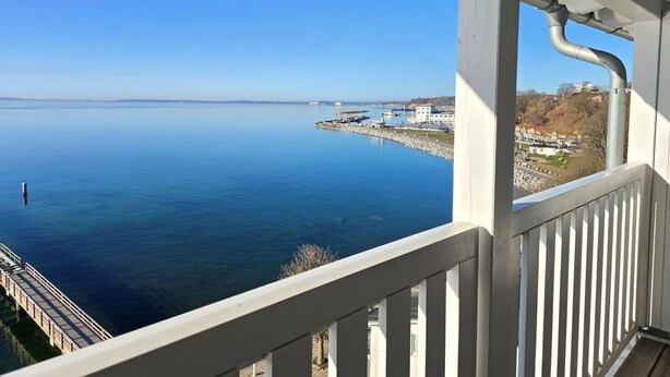 Vista desde un balcón sobre un cuerpo de agua tranquilo con un muelle que se adentra en él, edificios al fondo y un cielo despejado.