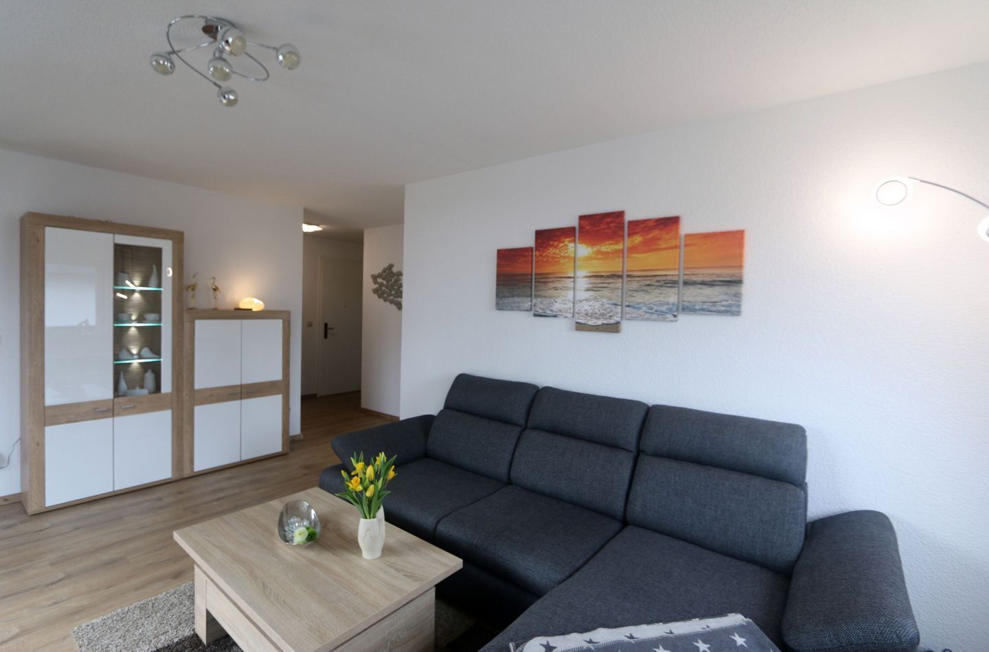 Una sala de estar moderna con un sofá seccional gris, una mesa de centro de madera con un jarrón de flores, un aparador con puertas de cristal y un cuadro colgado en la pared que representa un atardecer.