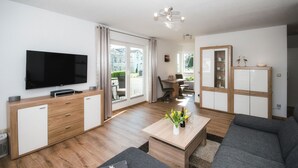 Een moderne woonkamer met een flatscreen televisie, houten meubels, een eethoek met tafel en stoelen, en uitzicht door een glazen deur.