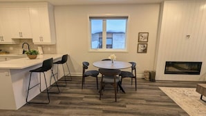 A modern kitchen with white cabinets, a dining area with a round table and chairs, a window with a view, and a fireplace. - Quiet 2BR Condo Near Hospital and Wi-Fi (Mount Forest)