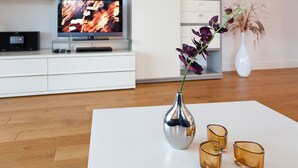 Un saló modern amb un televisor de pantalla plana, armaris blancs, una taula de cafè blanca, un vas amb flors morades i dos suporta-espelmes de vidre.