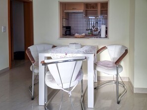 A dining area with a white table and chairs, a kitchen in the background, and a tiled floor. - Ferienhaus Zum Kreideblick, FeWo Muschel - Holiday home Zum Kreideblick, (Sassnitz auf Rügen)