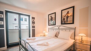 A neatly made double bed with white linens, two bedside lamps, and a view of a building through a glass door. - Villa Friede-Marie, App. Baltic Sea view - with fantastic sea view - Villa (Sassnitz auf Rügen)
