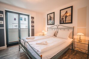 Ein ordentlich bezogenes Doppelbett mit weißen Bettwäsche, zwei Nachttische mit Lampen und Blick auf ein Gebäude durch eine Glastür.