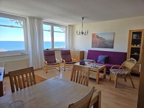 Ein Wohnzimmer mit Meerblick, einem Esstisch, Stühlen, einer Couch und einem Bücherregal.