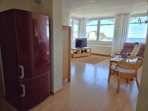 Ein Wohnzimmer mit Fernseher, Holzschrank, Korbstuhl und Blick aufs Meer.