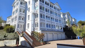 Un immeuble à plusieurs étages, avec un escalier menant à l’entrée, un balcon doté de balustrades en treillis et un espace de garage avec des portes fermées.