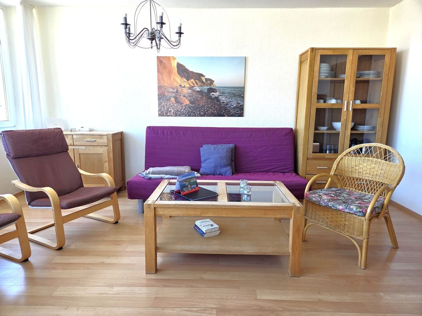 A living room with a purple sofa, a wooden coffee table, wicker chairs, a wooden cabinet with dishes, and a chandelier.