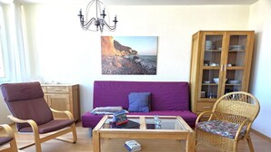 Un salon avec un canapé violet, une table basse en bois, des fauteuils en osier, un buffet en bois contenant de la vaisselle et un lustre.