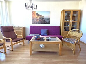 Ein Wohnzimmer mit einem violetten Sofa, einem Holztisch, Korbstühlen, einem Holzschrank mit Geschirr und einer Pendelleuchte.