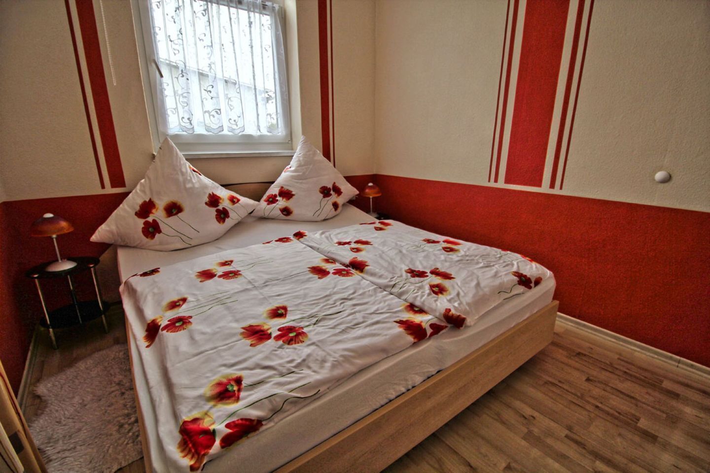 Ein Bett mit floralem Überwurf, zwei Kissen, eine Nachttischlampe und ein Fenster mit Aussicht.