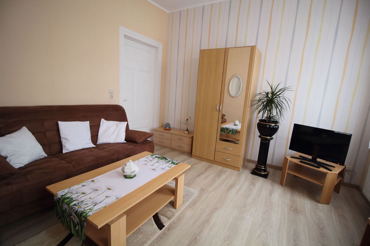 Ein Wohnzimmer mit braunem Sofa, einem Holztisch, einem Fernseher auf einem Schrank, einem Kleiderschrank und einem Topfpflanzen.