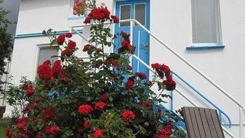 Ein weißes Gebäude mit blauer Tür und Fensterläden, eine Treppe mit blauen Geländern und ein Blumentopf mit roten Blüten.