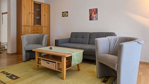 Ein Wohnzimmer mit grauem Sofa, zwei Sesseln, einem Holztisch und einem Schrank mit Glastüren.
