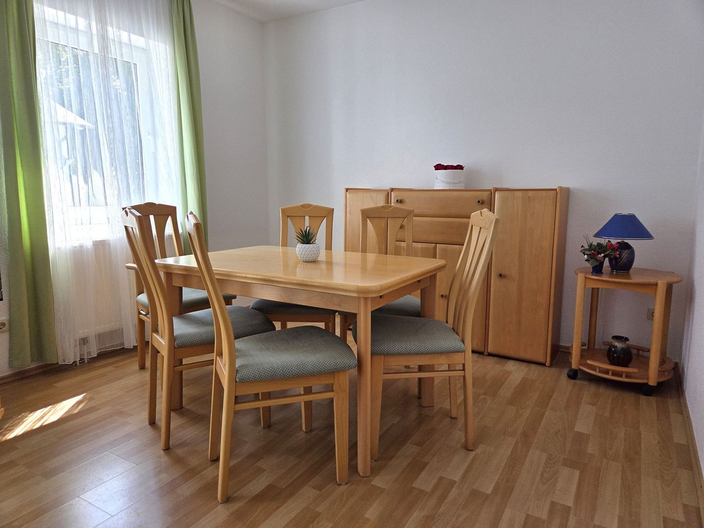Ein Essbereich mit einem Holztisch, Stühlen, einem kleinen Schrank und einem Beistelltisch mit einer Lampe.