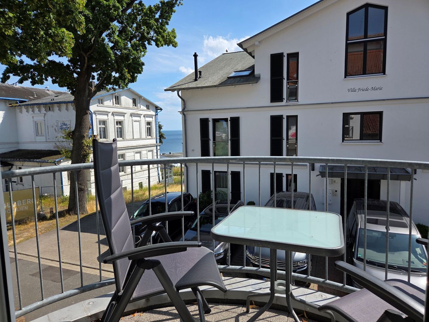 A balcony with a table and chairs, overlooking a street with parked cars and a building with a sign 'Villa Friede-Marie'.
