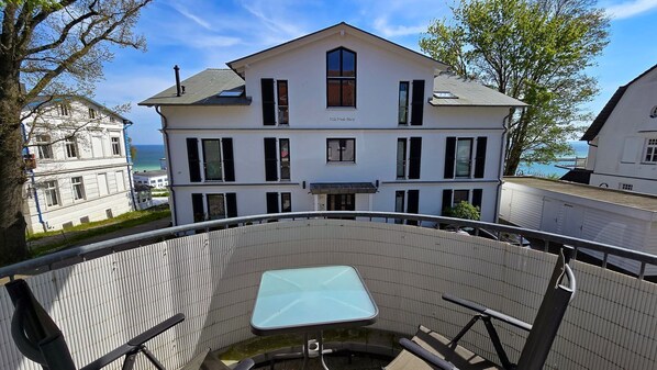 Ein Balkon mit Glastisch und Stühlen, mit Blick auf ein weißes Gebäude mit zahlreichen Fenstern und einem Balkon.
