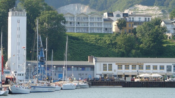 A marina with several boats docked, a white building with multiple windows and balconies, and a hillside with buildings in the background. - Villa Prinz Heinrich - App.3 with balcony and Baltic Sea view - Villa Prinz Heinrich, (Sassnitz auf Rügen)