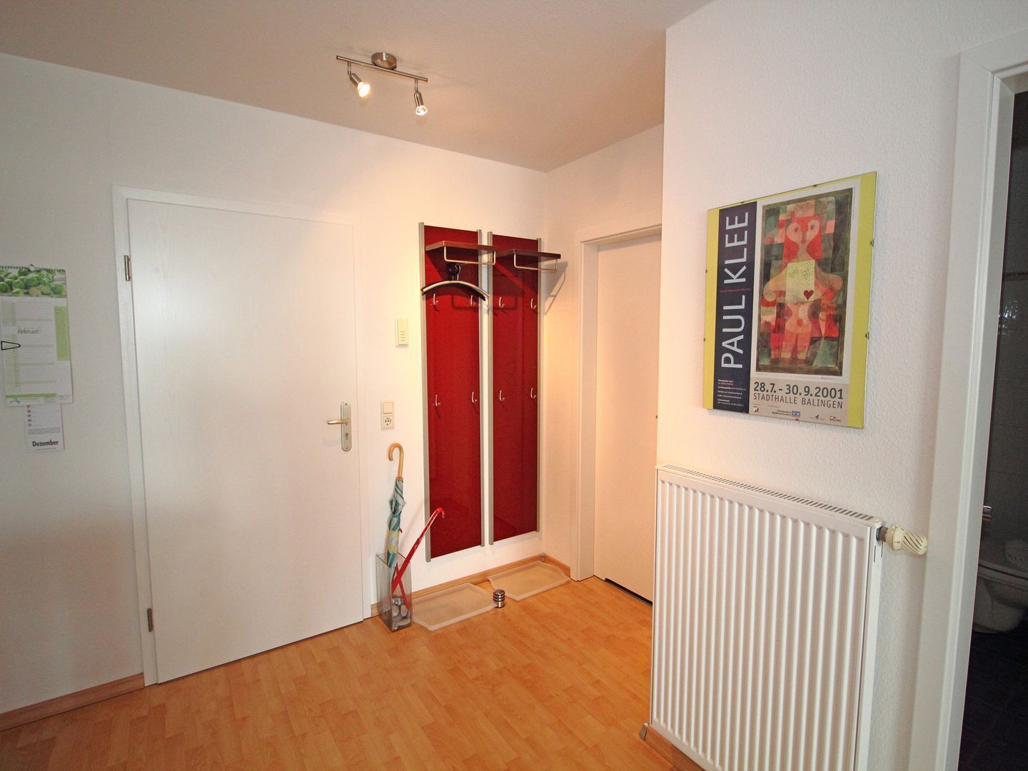 Un pasillo con suelo de madera, una puerta blanca, una puerta roja, un perchero, un cartel y un radiador.