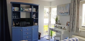 Ein Zimmer mit blauem Schrank, Schreibtisch, Stuhl, Bücherregal, Gemälde und einem Fenster mit Vorhängen.