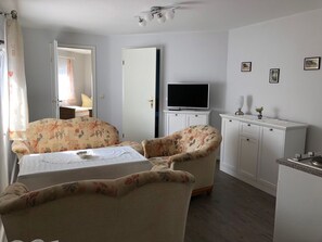 Una habitación con una cama, dos sofás de estampado floral, un televisor sobre un mueble y una pequeña cocina americana.