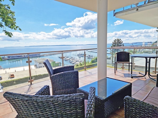 Ein Balkon mit Korbmöbeln, Glasgeländer und Blick auf eine Marina.