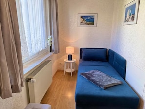 A small room with a blue sofa, a small table with a lamp, a radiator, and a window with curtains.