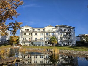 Ein mehrstöckiges Gebäude mit Balkonen, einem Teich mit glatter Wasseroberfläche und blauem Himmel.