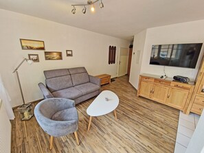 Ein Wohnzimmer mit einer Couch, zwei Sesseln, einem kleinen Tisch, einem Fernseher, Holzböden und einem Schrank.