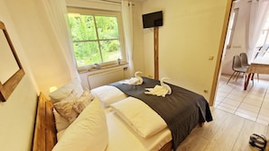 Una camera da letto con un letto, una TV, una finestra con tende, un pavimento in legno e un'area pranzo con sedie.