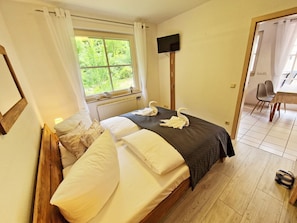 Ein Schlafzimmer mit Bett, Fernseher, Fenster mit Vorhängen, Holzboden und einer Essecke mit Stühlen.